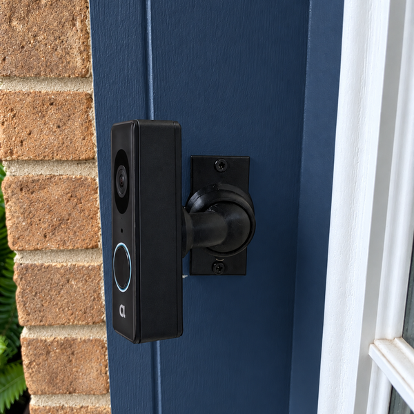Alula CAM-DB-JS1 on adjustable 15–90 degree swivel mount, left-facing view on navy blue painted trim beside warm tan brick